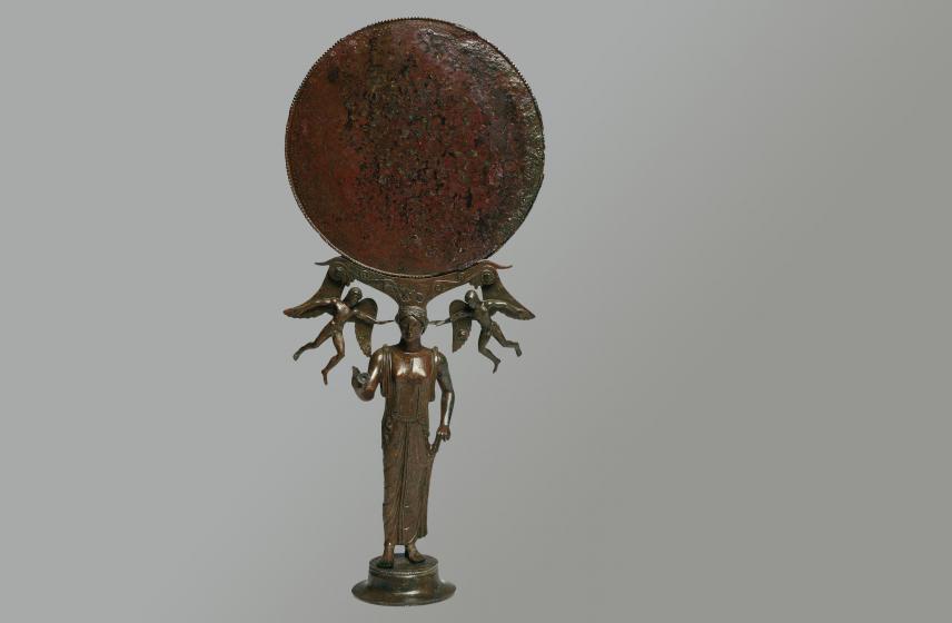 Ce beau miroir à pied évoque le goût des femmes grecques pour les objets de toilette et de parure. Le disque, autrefois réfléchissant, est bordé d'une double rangée d'oves et de perles et son support en volutes est orné d'élégants rinceaux incisés.